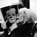 Black and white image of an elderly lady in a wheelchair holding up a photo of a baby close to her face