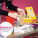 Woman handling a book and brown paper bag as though about to give to a customer amongst other photo books on a stall with the Impressions Gallery graphic for the event with details