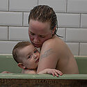 A mother and her child in a bathtub together, mother looking down and child directly at camera