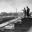 A black and white photograph of two men stood on a derelict building looking down on a landscape of a terrace of houses and factories in the background 