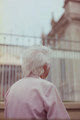 Elderly lady in pink checkered shirt with white hair, facing away from camera looking up at building 