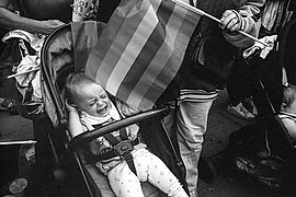 A black and white image of a baby crying in a pushchair with a flag above them in a crowd