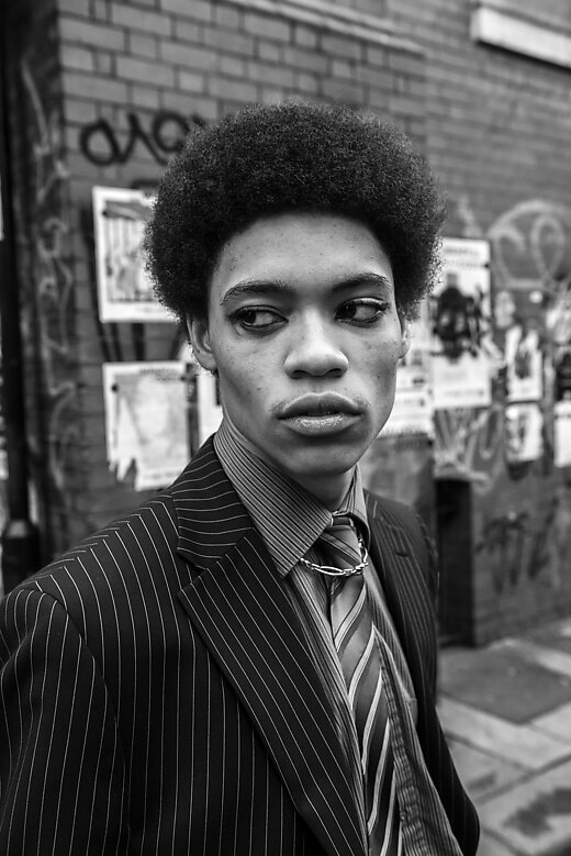 A man with dark eyeliner on and in a suit, close-up portrait, looking away from the camera in an urban setting 