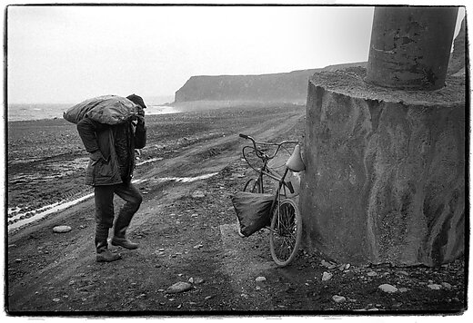 Mark Pinder - Scavenging coal on the beach at Easington Colliery, Co Durham, 1987 Black and white image of a man walking to his bike at the top of a beach. The bike is resting on a large concrete block and the man carries a large sack over his shoulder. It seems like quite a dreary day and the sand on the ground looks more like mud.